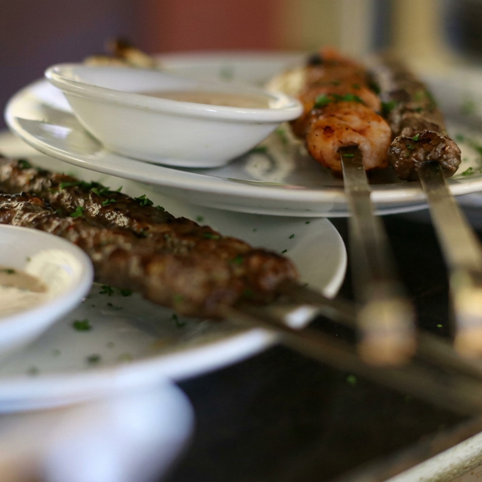 Grilled meat skewers and kebabs served on white plates with creamy dipping sauces and chopped herbs.
